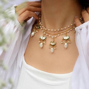 Peut inclure: Un collier doré à plusieurs rangs avec des perles et trois breloques pendantes. Les breloques présentent un motif floral vert et doré avec des perles pendantes. Le collier est porté sur un haut blanc.