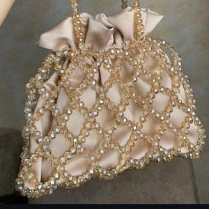 May include: A beige satin drawstring bag with a beaded overlay. The bag features a delicate pattern of clear, faceted beads in a diamond grid design. The handles are also beaded, and the bag is closed with a satin drawstring.