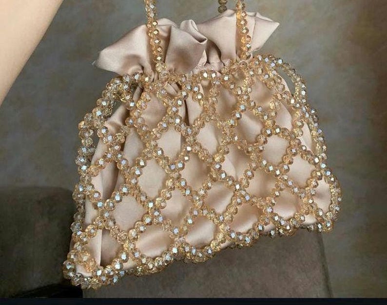 May include: A beige satin drawstring bag with a beaded overlay. The bag features a delicate pattern of clear, faceted beads in a diamond grid design. The handles are also beaded, and the bag is closed with a satin drawstring.