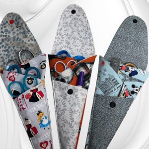 May include: Three elongated fabric scissor cases with snap closures. The left case features Alice in Wonderland characters, the center case has a skull and vine pattern, and the right case displays a Mickey Mouse and Paris-themed print.