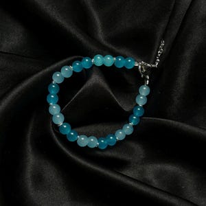 May include: A bracelet made of blue beads in varying shades, strung on a silver chain. The beads are round and glossy. The background is a black satin material. The bracelet is approximately 18 cm in length.