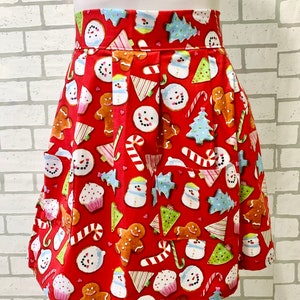 May include: Red apron with a Christmas cookie pattern. The pattern includes gingerbread men, snowmen, candy canes, cupcakes, and Christmas trees. The apron has two pockets.
