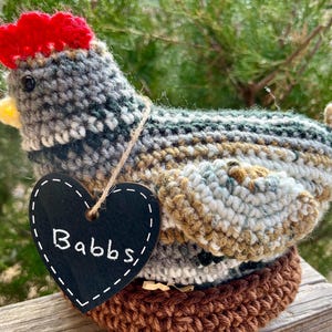May include: A handmade crochet chicken figurine with a red comb, yellow beak, and a heart-shaped tag that reads "Babbs". The chicken is perched on a brown crocheted nest. The body is a mix of gray, blue, and brown yarn.