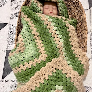May include: A baby swaddled in a handmade, green, and tan crochet blanket and hood, nestled in a woven basket. The blanket has a textured, geometric pattern. The basket is placed on a white and black patterned quilt.