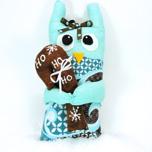 May include: A turquoise owl-shaped plush toy with large black and white eyes, a yellow beak, and a brown polka-dot bow. It holds a brown gingerbread cookie with "HO" text and a white snowflake. The owl is wrapped in patterned fabric.