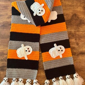 May include: A crocheted scarf featuring horizontal stripes in black, orange, and gray. The scarf is decorated with several white ghost appliqués, each with black eyes. The ends of the scarf are finished with multiple white yarn tassels.