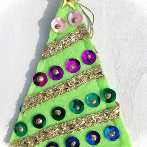 May include: A green felt Christmas tree ornament with gold glitter trim and colorful sequins. The tree has a gold star on top.