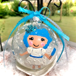 May include: A clear glass Christmas ornament with a painted image of a girl with blue hair and a white dress. The ornament is decorated with white snowflakes and glitter.
