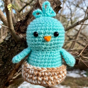 May include: A handmade crochet bird in a nest. The bird is light blue with black eyes and an orange beak. The nest is brown and white. The bird is perched on a tree branch against a blurred background of branches and sky.