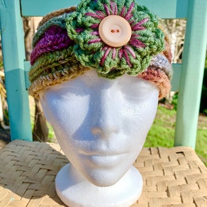 May include: A crocheted headband with a multi-colored striped pattern and a large green and purple flower with a button in the center.