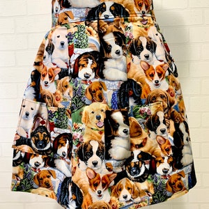 May include: A white apron with a colorful pattern of various dog breeds. The apron has two pockets on the front and a ruffled skirt.
