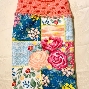 May include: A hanging kitchen towel with a coral crochet top and button fastening. The lower section has a patchwork design with floral patterns in pink, red, blue, and white. The towel is designed for hanging.