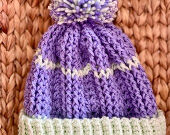 May include: A handmade crochet beanie in shades of purple and light green. The hat features a ribbed brim in light green, a body in purple with white accents, and a large pom-pom on top. The beanie is designed for warmth and style.