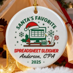 May include: White ceramic Christmas ornament with gold string. The ornament features the text "SANTA'S FAVORITE" and "SPREADSHEET SLEIGHER Anderson, CPA 2025" in red and green. A computer graphic with a Santa hat is in the center.
