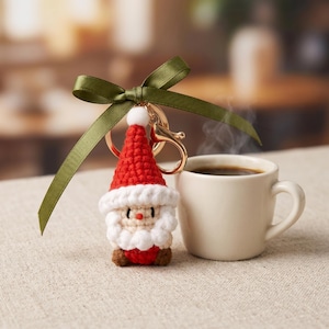May include: A handmade crochet Santa Claus keychain with a red hat, white beard, and brown boots. The keychain is attached to a gold-colored clasp and a green ribbon. A white mug of coffee sits next to the keychain on a neutral surface.