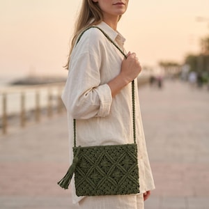 May include: An olive green macrame crossbody bag featuring a diamond-patterned knot design and a matching side tassel. The bag is suspended by a long, braided cord strap, showcasing intricate hand-knotted details and a rectangular silhouette.
