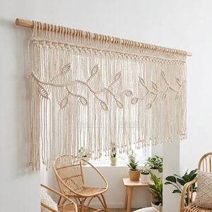 Pode incluir: Uma tapeçaria de macramé cor de creme com um padrão de folhas criado através de intrincados desenhos de nós. A peça está suspensa numa vara de madeira lisa de tom claro, com longas franjas soltas que pendem da extremidade inferior.