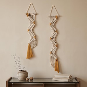 May include: Two vertical wall hangings made of off-white macrame cord with mustard yellow accents. Each piece features a zigzag pattern suspended from a wooden dowel with a triangular hanging cord and a long mustard yellow tassel at the bottom.