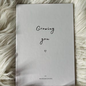 Könnte beinhalten: Ein weißes, rechteckiges Buch mit schwarzem Text auf dem Einband, der Growing you lautet, gefolgt von einem kleinen Herzsymbol und dem Text BY MAMAMADEIRL am unteren Rand. Der Einband hat eine glatte, matte Oberfläche und ein klares, zentriertes Design.