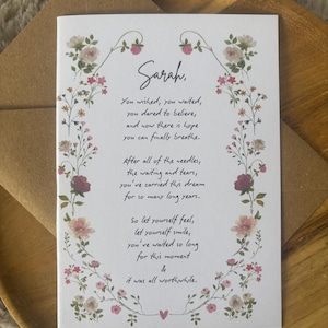 May include: A white greeting card with a floral border of pink, red, and white flowers. The text reads: Sarah, You wished, you waited, you dared to believe, and now there is hope you can finally breathe. After all of the needles, the waiting and tears, you've carried this dream for so many long years. So let yourself feel, let yourself smile, you've waited so long for this moment & it was all worthwhile.