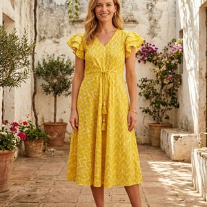 May include: A yellow midi dress with a white leaf pattern, V-neck, and ruffled sleeves. The dress has a tie waist with tassels and is worn with brown sandals. The background features potted plants and a stone walkway.