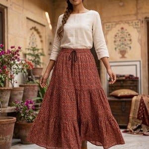 May include: A woman in a cream-coloured long-sleeved top and a tiered, rust-coloured skirt with a floral pattern. The skirt has a drawstring waist. She is wearing sandals. The background includes potted plants and a decorative wall.
