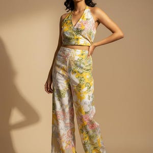 Könnte beinhalten: Ein zweiteiliges Outfit mit floralem Print, bestehend aus einem Neckholder-Top und einer Hose mit weitem Bein. Das Oberteil und die Hose haben eine weiße Basis mit gelben, rosa und grauen Blumenmustern. Das Outfit wird mit beigen Sandalen getragen.