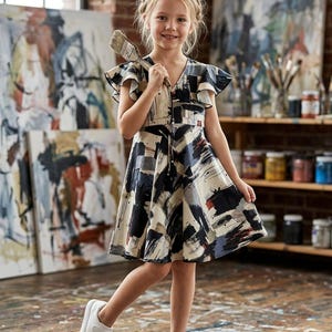 Op de afbeelding: Een jong meisje draagt een jurk met patroon en witte sneakers, met een penseel in de hand. De jurk heeft een abstract ontwerp in zwart, wit en blauw. De achtergrond toont schilderijen, kunstbenodigdheden en een bakstenen muur.
