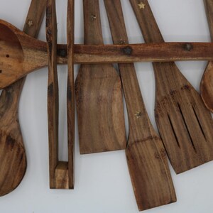 May include: A collection of wooden kitchen utensils, including spoons, spatulas, and tongs. The utensils are made of a light brown wood and have a natural grain pattern. Some utensils have small star-shaped accents. The set is arranged on a white background.