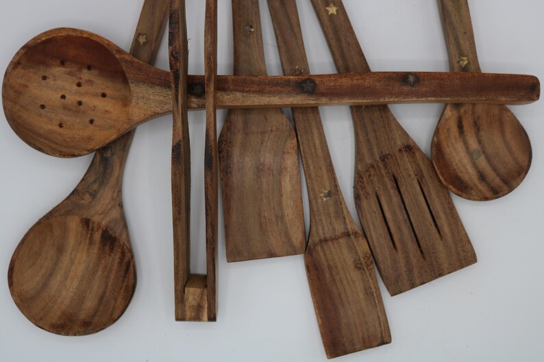 May include: A collection of wooden kitchen utensils, including spoons, spatulas, and tongs. The utensils are made of a light brown wood and have a natural grain pattern. Some utensils have small star-shaped accents. The set is arranged on a white background.