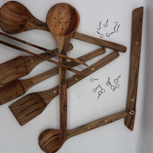 May include: Wooden kitchen utensils, including a slotted spoon, spatula, and tongs, are arranged with a wooden hanging rack. The utensils are a warm brown color and have small star-shaped accents. Small metal hooks are also included.