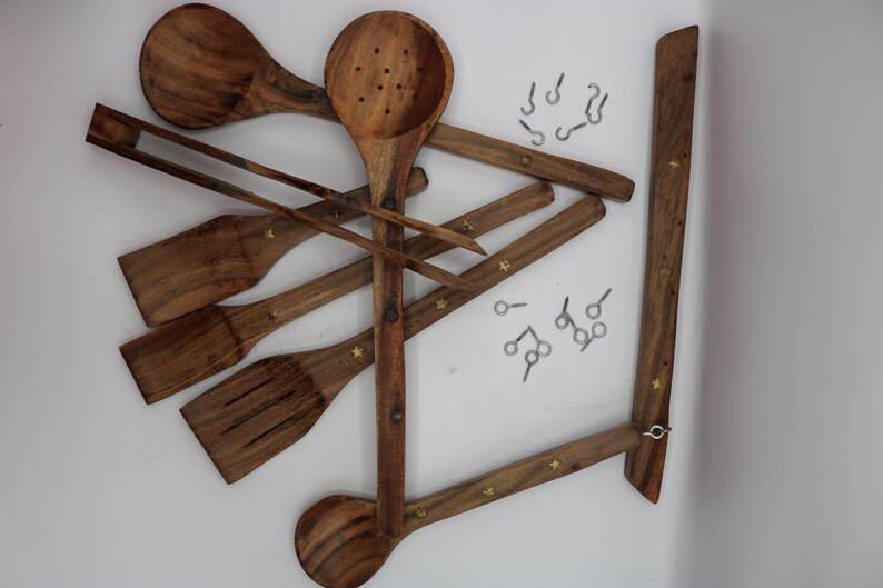 May include: Wooden kitchen utensils, including a slotted spoon, spatula, and tongs, are arranged with a wooden hanging rack. The utensils are a warm brown color and have small star-shaped accents. Small metal hooks are also included.
