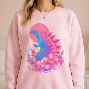 Può includere: Felpa rosa chiaro con un design colorato di Godzilla, un sole rosa e dettagli floreali. Il Godzilla è raffigurato in tonalità blu, viola e arancioni. I fiori sono rosa e bianchi. La felpa è a girocollo.