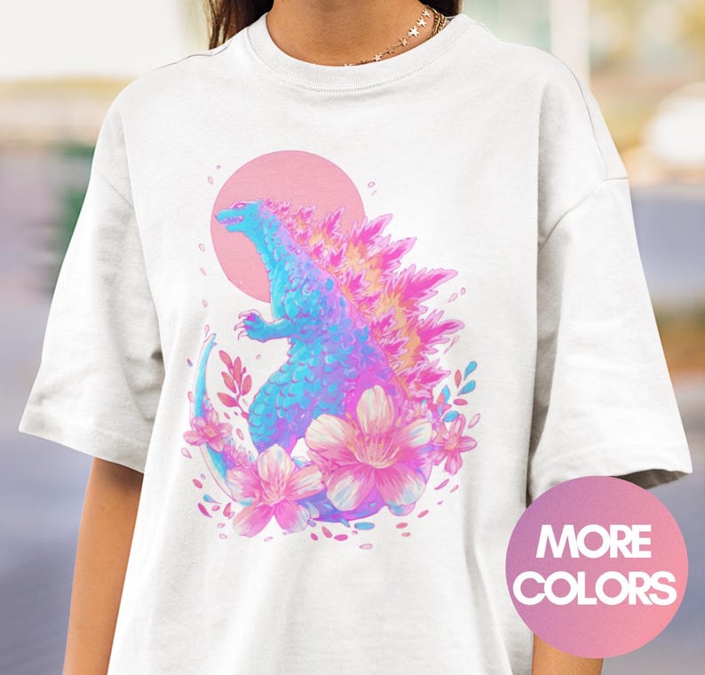 Peut inclure: T-shirt blanc avec une illustration color&eacute;e d'une cr&eacute;ature ressemblant &agrave; Godzilla, avec un soleil rose en arri&egrave;re-plan. La cr&eacute;ature est bleue et rose, entour&eacute;e de fleurs roses et de feuilles. Le t-shirt a un col rond et des manches courtes.