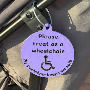 Peut inclure: Une étiquette ronde violet clair attachée au cadre d'une poussette avec un câble métallique. L'étiquette porte le texte noir : Please treat as a wheelchair. My pushchair keeps me safe. Un symbole de fauteuil roulant noir est centré.