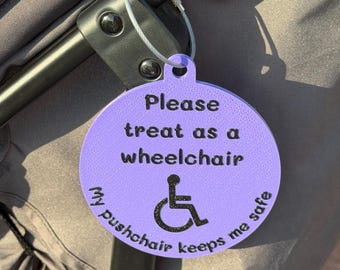 Puede incluir: Una etiqueta redonda de color morado claro sujeta al marco de un cochecito con un cable metálico. La etiqueta tiene texto negro que dice: Please treat as a wheelchair. My pushchair keeps me safe. Un símbolo de silla de ruedas negro.