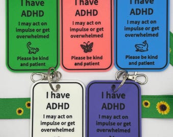 Puede incluir: Cinco etiquetas rectangulares de plástico en verde, coral, azul, blanco y morado, cada una con un cierre de mosquetón plateado. El texto dice: I have ADHD, I may act on impulse or get overwhelmed, Please be kind and patient. Cada etiqueta presenta un pequeño icono.