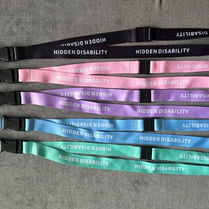 Puede incluir: Cinco cordones en negro, rosa, morado, azul y verde menta. Cada uno presenta el texto blanco HIDDEN DISABILITY repetido a lo largo de la cinta, una hebilla de seguridad de pl&aacute;stico negro y un mosquet&oacute;n de metal plateado con un peque&ntilde;o coraz&oacute;n rosa.