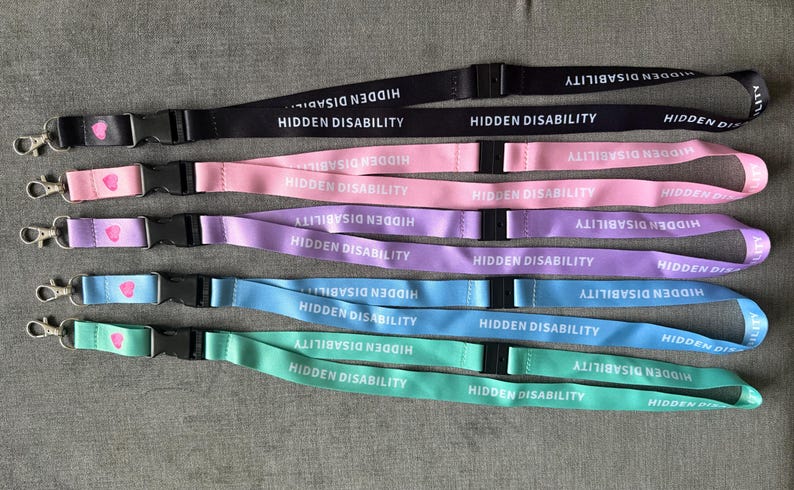 Puede incluir: Cinco cordones en negro, rosa, morado, azul y verde menta. Cada uno presenta el texto blanco HIDDEN DISABILITY repetido a lo largo de la cinta, una hebilla de seguridad de pl&aacute;stico negro y un mosquet&oacute;n de metal plateado con un peque&ntilde;o coraz&oacute;n rosa.