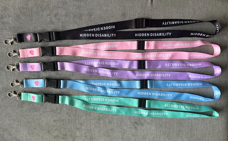 May include: Five lanyards in black, pink, purple, blue, and mint green. Each features the white text HIDDEN DISABILITY repeated along the strap, a black plastic safety breakaway buckle, and a silver metal lobster clasp with a small pink heart icon.