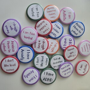 May include: A collection of colorful round buttons with various phrases related to neurodiversity and mental health. The buttons feature text like "I am Non Verbal," "I have Anxiety," and "I am Autistic." The buttons are in shades of pink, blue, green, and orange.