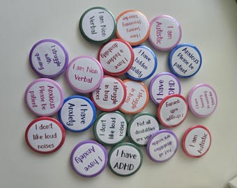 May include: A collection of colorful round buttons with various phrases related to neurodiversity and mental health. The buttons feature text like "I am Non Verbal," "I have Anxiety," and "I am Autistic." The buttons are in shades of pink, blue, green, and orange.