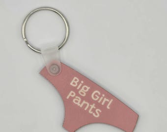 May include: A pink keychain with a silver ring and a clear plastic strap. The keychain is shaped like a pair of underwear and has the words "Big Girl Pants" printed on it in white.