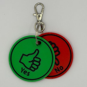 May include: A keychain featuring two circular tokens. One is green with a black thumbs-up and the word "Yes." The other is red with a black thumbs-down and the word "No." The keychain has a silver clasp.