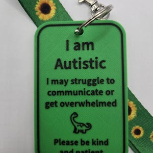 Peut inclure: Une étiquette rectangulaire en plastique vert avec une bordure noire et le texte noir I am Autistic, I may struggle to communicate or get overwhelmed, et Please be kind and patient, avec une icône de dinosaure et un cordon tour de cou tournesol.