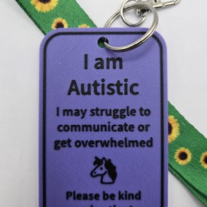 Puede incluir: Una etiqueta rectangular morada con texto negro que dice I am Autistic, I may struggle to communicate or get overwhelmed, y Please be kind and patient, con un icono de unicornio. Est&aacute; unida a un cord&oacute;n verde con estampado de girasoles.