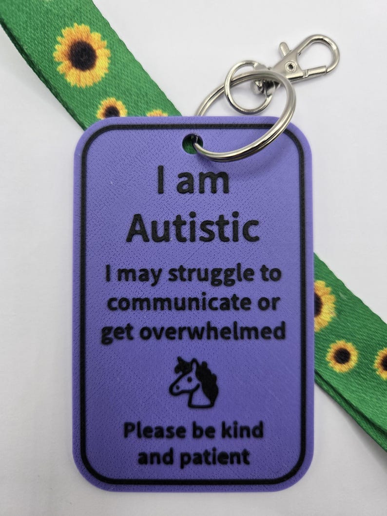 Puede incluir: Una etiqueta rectangular morada con texto negro que dice I am Autistic, I may struggle to communicate or get overwhelmed, y Please be kind and patient, con un icono de unicornio. Est&aacute; unida a un cord&oacute;n verde con estampado de girasoles.