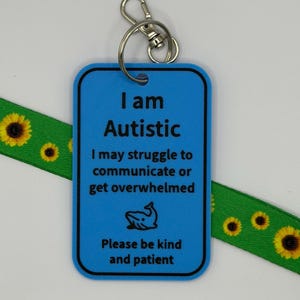 Puede incluir: Una etiqueta rectangular de pl&aacute;stico azul con un llavero de metal, que presenta el texto en negro: I am Autistic, I may struggle to communicate or get overwhelmed, un icono de ballena y Please be kind and patient, sobre un cord&oacute;n verde con girasoles.
