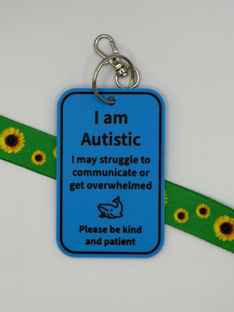 Puede incluir: Una etiqueta rectangular de pl&aacute;stico azul con un llavero de metal, que presenta el texto en negro: I am Autistic, I may struggle to communicate or get overwhelmed, un icono de ballena y Please be kind and patient, sobre un cord&oacute;n verde con girasoles.