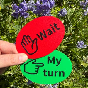 Peut inclure: Deux panneaux ovales, un rouge et un vert, tenus devant un fond floral. Le panneau rouge indique "Wait" avec un symbole de main. Le panneau vert indique "My turn" avec un symbole de main pointant.
